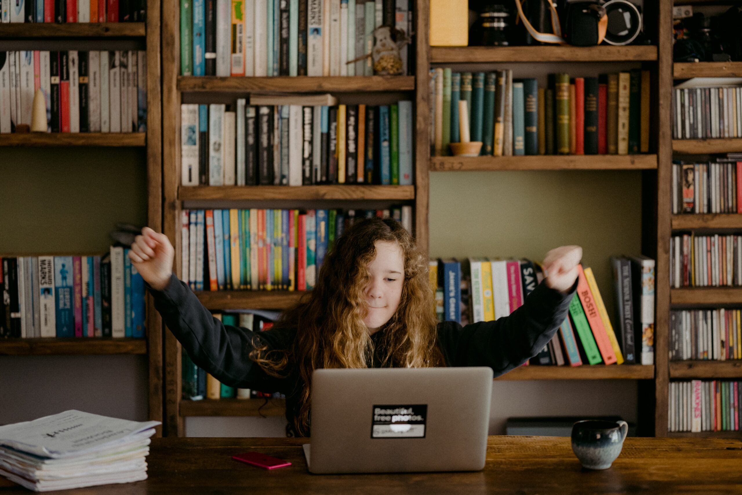 Kid raising her arms in celebration while looking at laptop in front of bookshelf.
