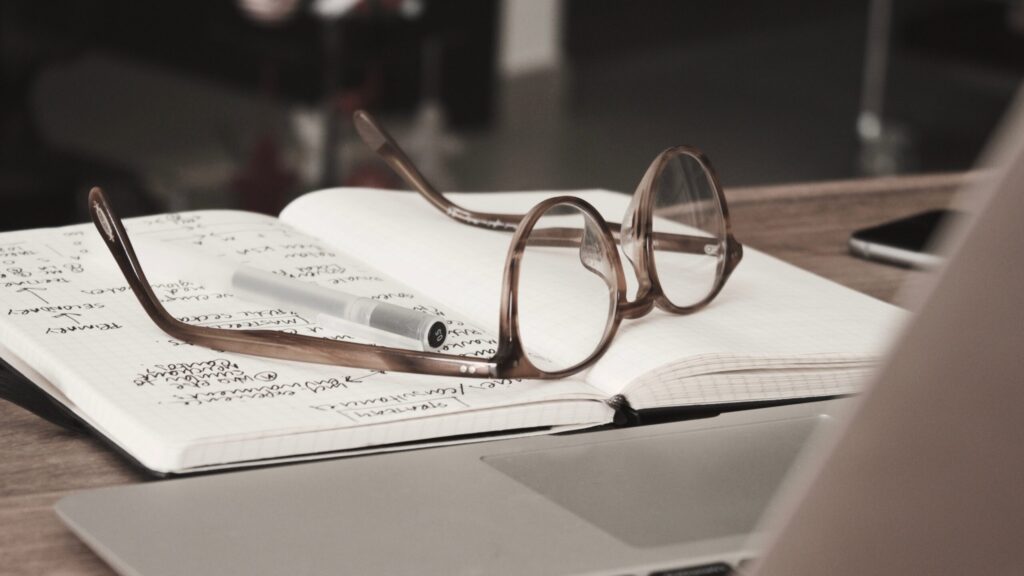 Glass laying on an open notebook on a table next to an open laptop.