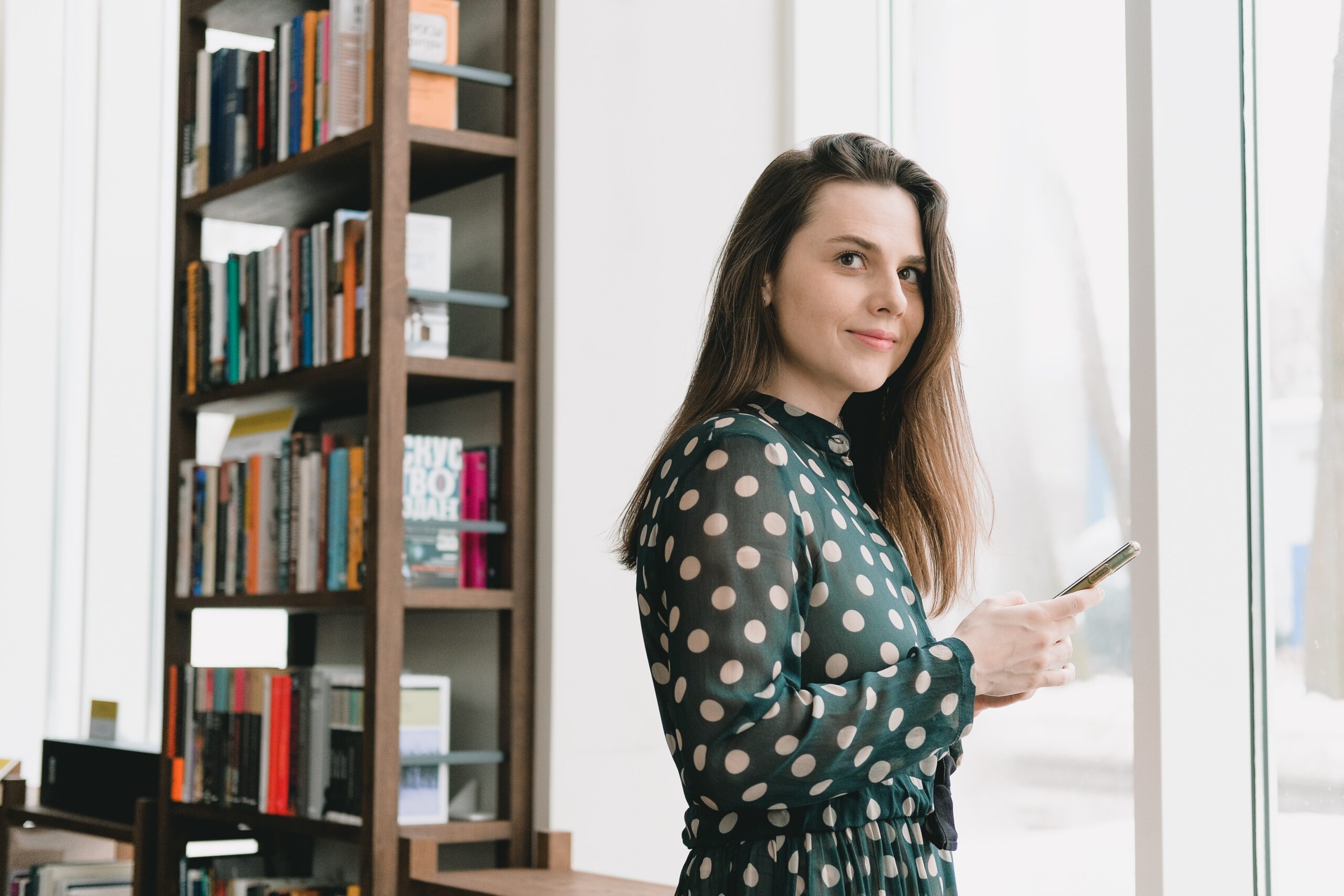 Woman staring out window, phone in hand, with bookshelf behind her.