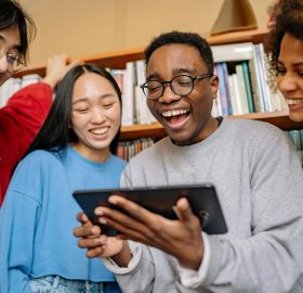 Four people excitedly huddled around an iPad in front of a bookcase.